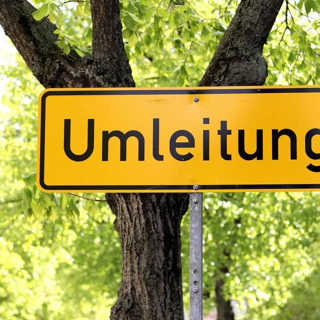 Ein Straßenschild aus Deutschland, das im Hintergrund gelb ist und die schwarze Aufschrift "Umleitung" trägt, steht vor einer Straßenkulisse mit einer begrünten Allee.