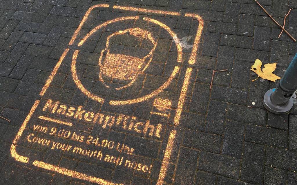 Ein Graffiti auf dem Pflaster der Fußgängerzone weist auf die Maskenpflicht hin.
