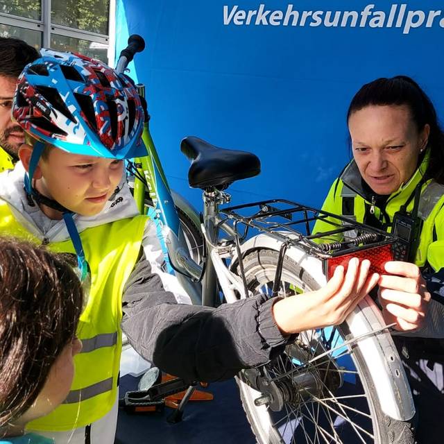 Ein Junge mit gelber Warnweste hält eine Rücklampe an ein Fahrrad. Daneben steht eine Polizistin.