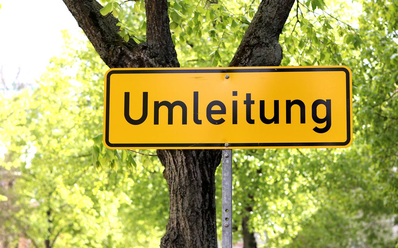 Ein Straßenschild aus Deutschland, das im Hintergrund gelb ist und die schwarze Aufschrift "Umleitung" trägt, steht vor einer Straßenkulisse mit einer begrünten Allee.