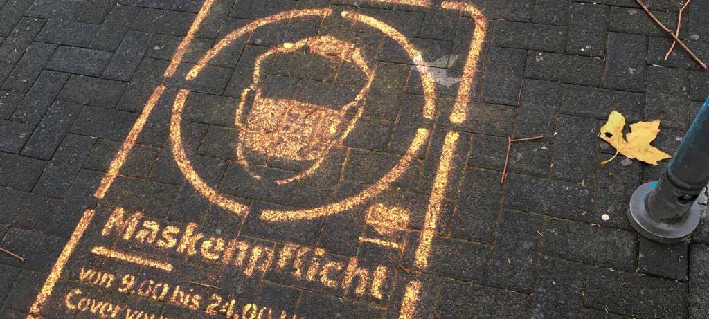 Ein Graffiti auf dem Pflaster der Fußgängerzone weist auf die Maskenpflicht hin.