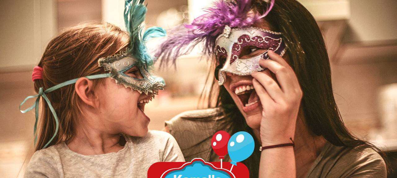 Eine Mutter lächelt ihr Kind an, während beide eine Karnevalsmaske tragen und zu Hause sind.