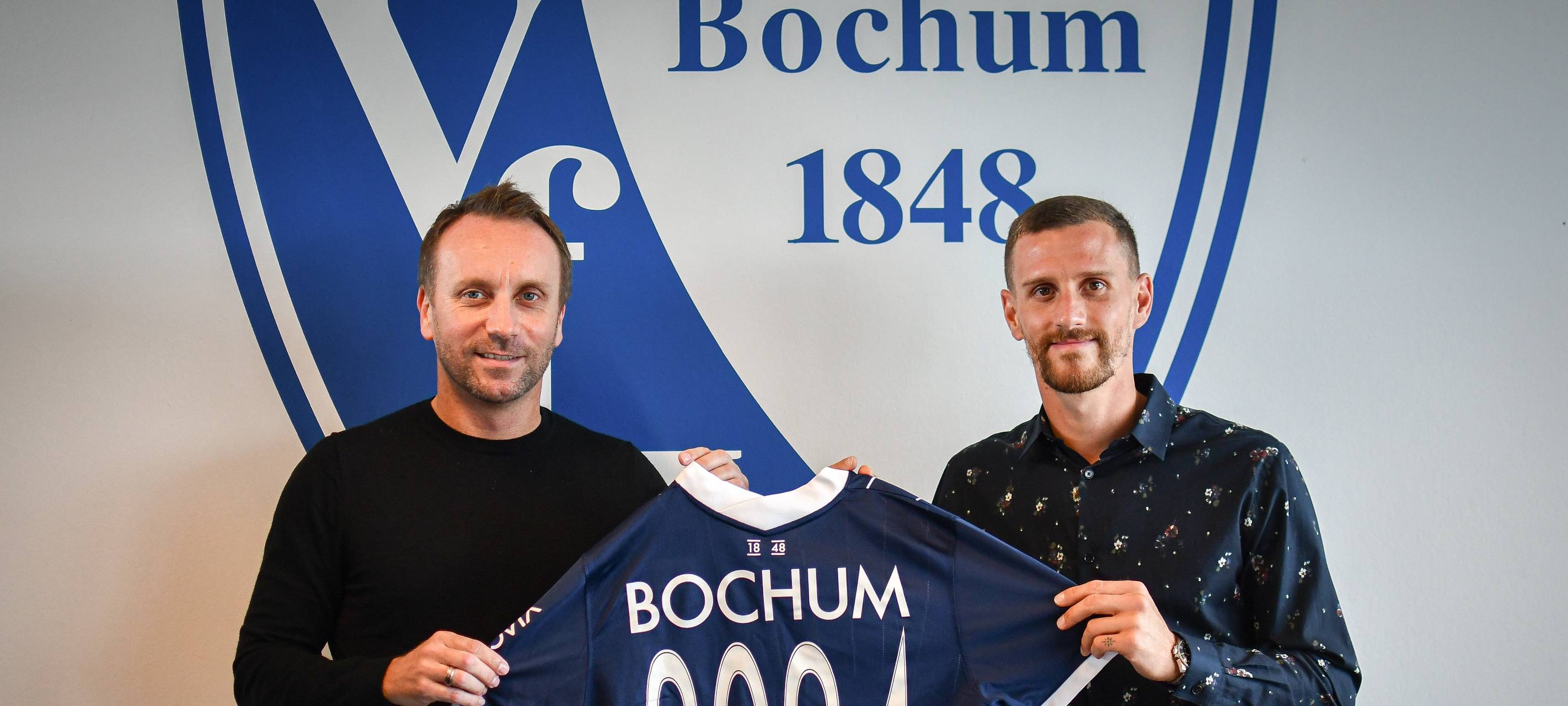 Zwei Männer lächeln in die Kamera und halten ein VfL-Trikot in den Händen.