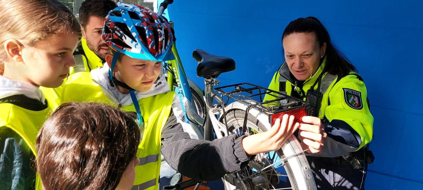Ein Junge mit gelber Warnweste hält eine Rücklampe an ein Fahrrad. Daneben steht eine Polizistin.