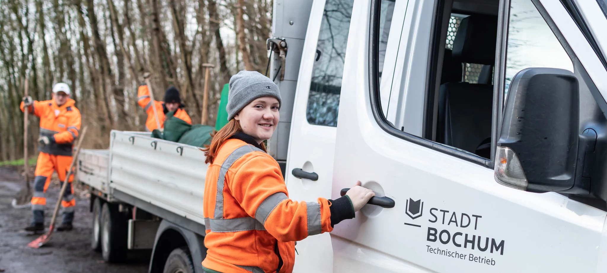 Junge Frau im orangenen Arbeitsanzug steht an einem Auto und lächelt in die Kamera.